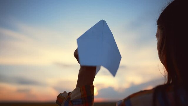 View from back on hand of young girl who holds paper airplane and launches it forward into field at sunset. Outdoor games with fly toy at leisure activity. Sporty lifestyle in summer. Push with force.
