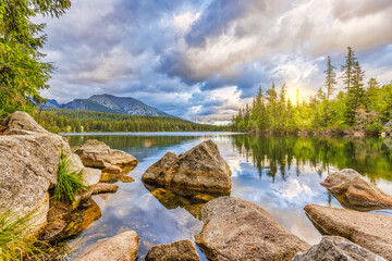 Beautiful mountain landscape, picturesque High Tatra lake in summer morning. Amazing nature panorama, rocks boulders with calm water reflection and pine tree forest. Tranquil scenic view, sunny clouds