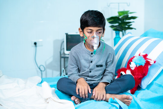 Worried sick kid with oxygen mask sitting on bed at hospital ward - concept of breathing problems, respiratory infection and health care treatment.