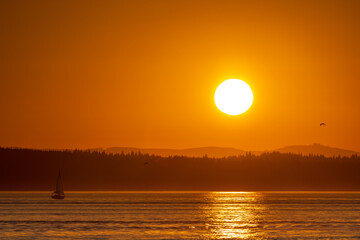 A brilliant orange sun sets over Puget Sound with silhouetted hills in the background and reflections on the ocean in the foreground