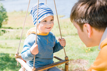 Stylish young father swings his happy three-year-old son dressed in yellow blue clothes on a wooden swing on a sunny day in the countryside. The concept of child care, safety for children.