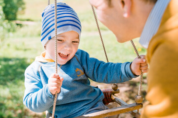 Stylish young father swings his happy three-year-old son dressed in yellow blue clothes on a wooden swing on a sunny day in the countryside. The concept of child care, safety for children. - obrazy, fototapety, plakaty