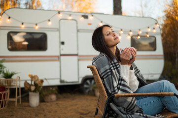 Beautiful caucasian woman is sitting wrapped in a plaid and drinking a warming drink outdoors. Traveling in a motor home in autumn.