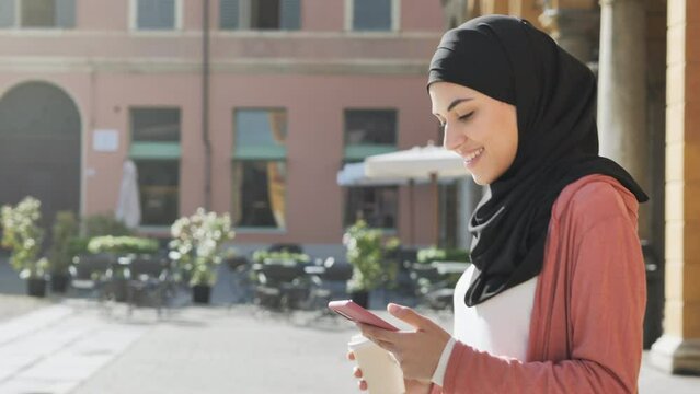 muslim female using smart phone mobile walks in european city square,pleased arab girl wearing hijab uses smartphone while walking tracking shot steadicam slow motion side view