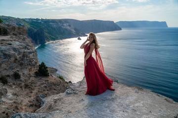 A girl with flowing hair in a long red dress stands on a rock above the sea. The stone can be seen in the sea. Sunny path to the sea from the sun.