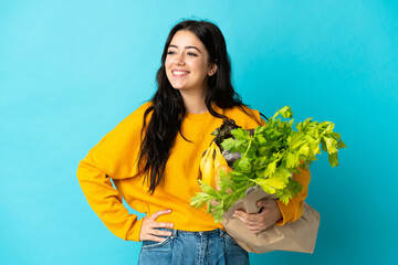 Young woman holding a grocery shopping bag isolated on blue background posing with arms at hip and...