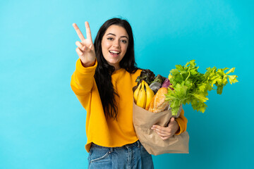 Young woman holding a grocery shopping bag isolated on blue background smiling and showing victory sign