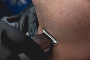 A barber uses a t-blade hair clipper to shave the facial hair and beard below the chin of a...