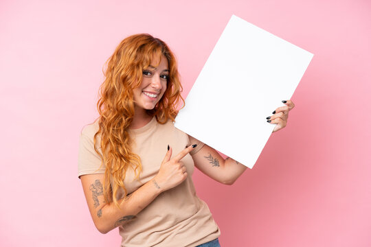 Young caucasian woman isolated on pink background holding an empty placard with happy expression and pointing it