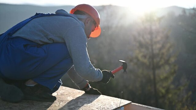 Carpenter hammering nail into OSB panel on the roof top of future cottage in the evening. Man worker building wooden frame house. Carpentry and construction concept.