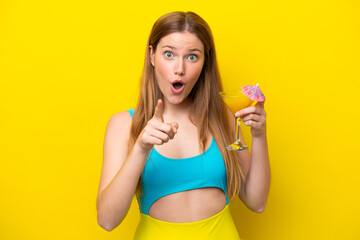 Young caucasian woman holding a cocktail isolated on yellow background surprised and pointing front