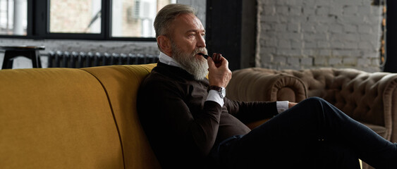 Senior thoughtful man smoking tobacco pipe on sofa