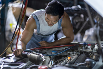 Asian car mechanic looks at the engine with a loud noise and checks the engine in the garage.