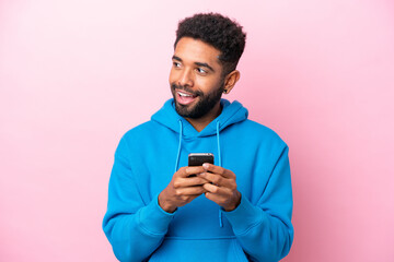 Young Brazilian man isolated on pink background using mobile phone and looking up