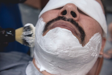 A barber uses a neck duster brush to apply shaving cream to the chin and sideburns of a client at a...