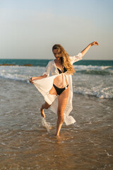 Chica joven con pelo largo en bikini en la orilla de la playa jugando con el agua del mar