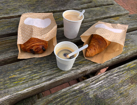 Paper Cups With Tasty Coffee And French Croissant In The Paper Package, Takeaway Morning Meal Snack-to-go On The Old Wooden Table Outside In The Park, Top View