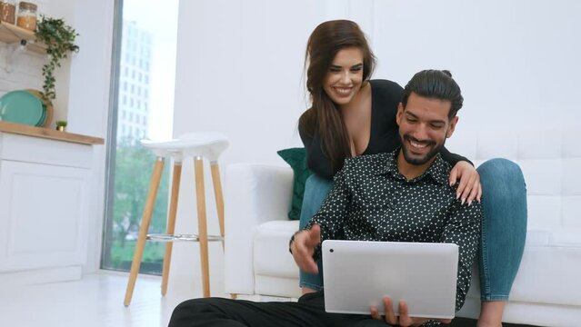 Young couple relaxing in living room together looking at laptop. White woman smiling sitting on couch leaning on her partner sitting on the floor. Indoor shot. High quality 4k footage