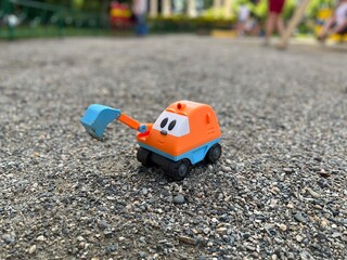 A little bright orange children's toy excavator explores the gravel surface. An excavator pulled down its bucket