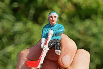 a hand holds one small plastic toy figurine of a blue hockey player outdoors on a green background - Powered by Adobe