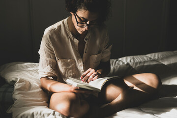 Young woman reading in her room with the light from the window sitting on the bed. Woman spending her leisure time reading book at night before sleeping. Girl enjoying her reading time - obrazy, fototapety, plakaty