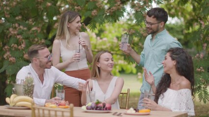 Group of happy young people cheering with drinks and eating fruits by the pool in the garden