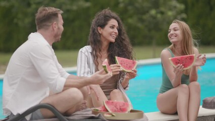 Group of young people standing by the swimming pool and eating watermelon in the house backyard