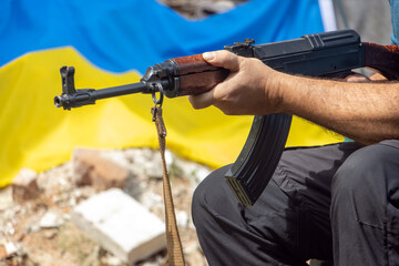 A man with an assault rifle sits in front of ruins with a Ukrainian flag