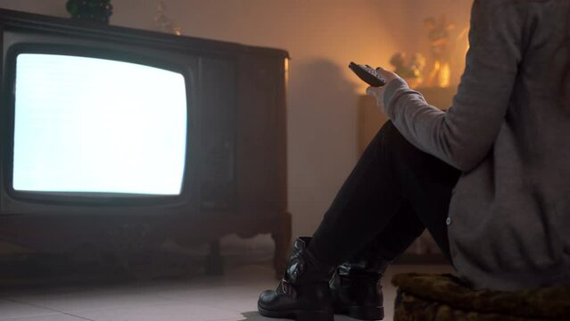 Dolly shot of woman sitting on the floor in old dark illuminated room and changing TV channels with remote control, female searching broadcast programs for home entertainment. Old broken vintage