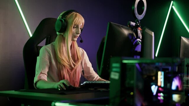 Young gamer girl wearing pink clothes and headset sitting in gaming chair by a desk focused on playing an online game. Blurred foreground. High quality 4k footage