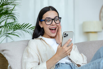 A beautiful woman in glasses is happy she received good news online message, she is delighted sitting on the sofa at home