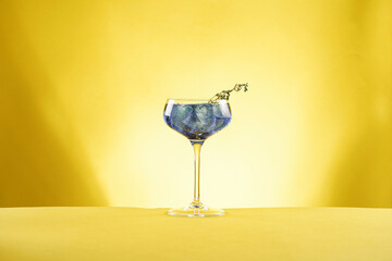 Dark blue drink in a vintage glass for sparkling wine - pea flower tea or blue curacao sirup cocktail with thyme branches on yellow backgroud. Copy space for text
