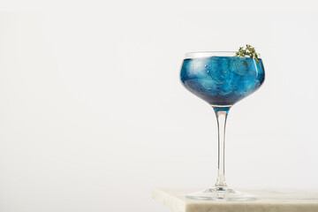 Dark blue drink in a vintage glass for sparkling wine - pea flower tea or blue curacao sirup cocktail with thyme branches on white backgroud. Copy space for text