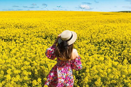Beautiful girl with dress and hat in the Canola Fields. Blooming canola fields under a blue sky with clouds. Beautiful yellow flowers. Happy girl in nature.
