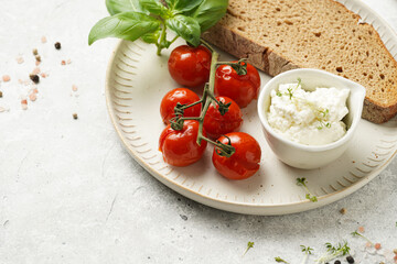 Roasted cherry tomatoes with branch, fresh cottage cheese, green basil and a slice of whole wheat bread on a plate on grey background