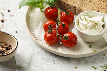 Roasted cherry tomatoes with branch, fresh cottage cheese, green basil and a slice of whole wheat bread on a plate on grey background