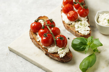 A sandwich with roasted cherry tomatoes with branch, fresh cottage cheese, green basil on a slice of whole wheat bread on a marble board on grey background