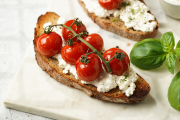 A sandwich with roasted cherry tomatoes with branch, fresh cottage cheese, green basil on a slice of whole wheat bread on a marble board on grey background