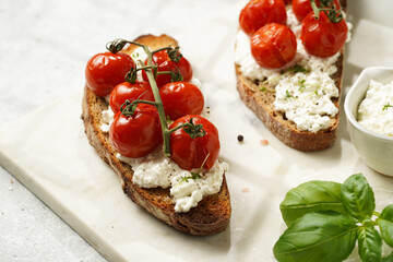 A sandwich with roasted cherry tomatoes with branch, fresh cottage cheese, green basil on a slice of whole wheat bread on a marble board on grey background