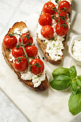 A sandwich with roasted cherry tomatoes with branch, fresh cottage cheese, green basil on a slice of whole wheat bread on a marble board on grey background