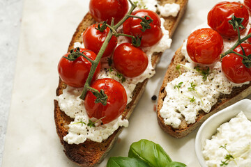 A sandwich with roasted cherry tomatoes with branch, fresh cottage cheese, green basil on a slice of whole wheat bread on a marble board on grey background