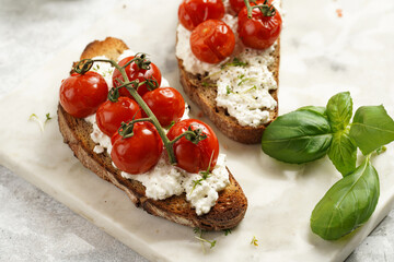 A sandwich with roasted cherry tomatoes with branch, fresh cottage cheese, green basil on a slice of whole wheat bread on a marble board on grey background