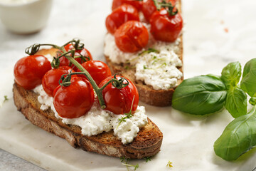 A sandwich with roasted cherry tomatoes with branch, fresh cottage cheese, green basil on a slice of whole wheat bread on a marble board on grey background