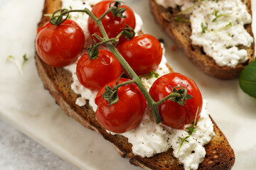 A sandwich with roasted cherry tomatoes with branch, fresh cottage cheese, green basil on a slice of whole wheat bread on a marble board on grey background