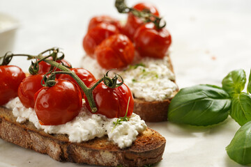 A sandwich with roasted cherry tomatoes with branch, fresh cottage cheese, green basil on a slice of whole wheat bread on a marble board on grey background