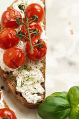 A sandwich with roasted cherry tomatoes with branch, fresh cottage cheese, green basil on a slice of whole wheat bread on a marble board on grey background