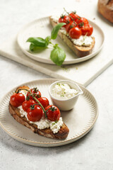 A sandwich with roasted cherry tomatoes with branch, fresh cottage cheese, green basil on a slice of whole wheat bread on a round plate on grey background