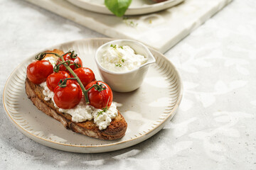 A sandwich with roasted cherry tomatoes with branch, fresh cottage cheese, green basil on a slice of whole wheat bread on a round plate on grey background