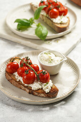 A sandwich with roasted cherry tomatoes with branch, fresh cottage cheese, green basil on a slice of whole wheat bread on a round plate on grey background
