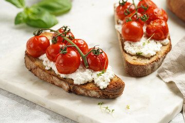A sandwich with roasted cherry tomatoes with branch, fresh cottage cheese, green basil on a slice of whole wheat bread on a marble tray on grey background